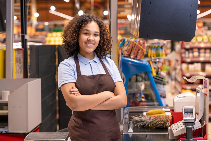 Pretty female in apron standing by cashbox in supermarket and crossing arms by chest on background of shelves with food products